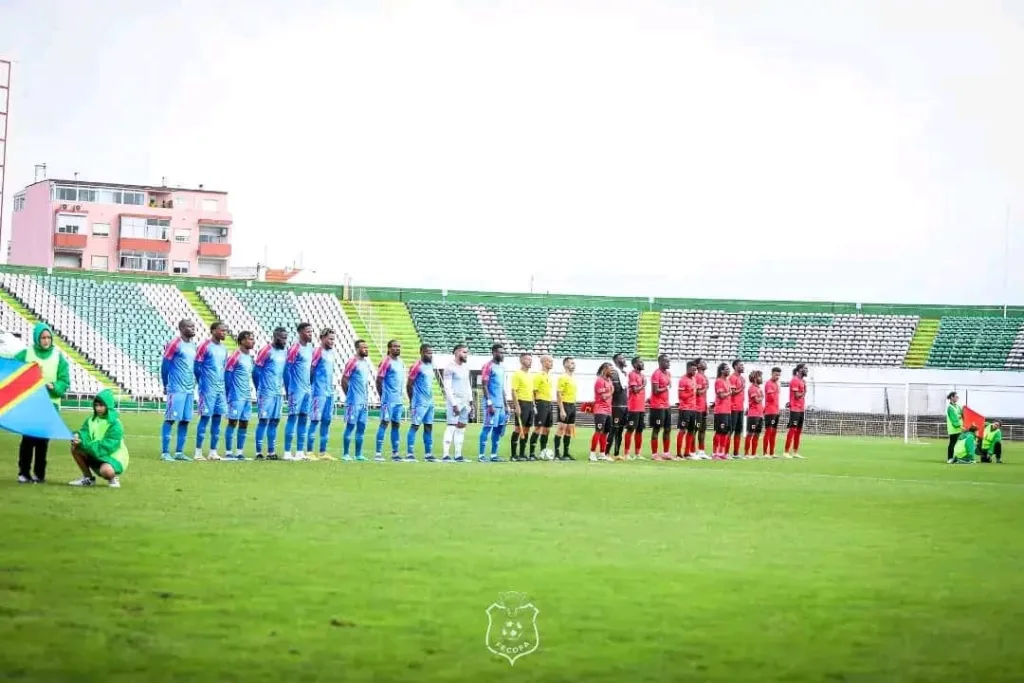 Sur la photographie, deux équipes de football dynamiques s'affrontent sur un terrain bien entretenu. À gauche, les joueurs vêtus du maillot bleu ciel de la RDC pressent vigoureusement vers l'avant, tandis qu'à droite, l'équipe de l'Angola, habillée en rouge et noir, forme un mur défensif, attendant l'attaque imminente. Le stade, plein à craquer, montre une mer de fans enthousiastes, leurs visages peints avec les drapeaux de leurs pays respectifs. En arrière-plan, le ciel d'après-midi est teinté de nuances dorées et orangées, signalant le coucher du soleil. L'intensité sur le visage des joueurs, couplée à l'atmosphère énergique, encapsule l'esprit de rivalité amicale, d'unité et de passion pour le sport.