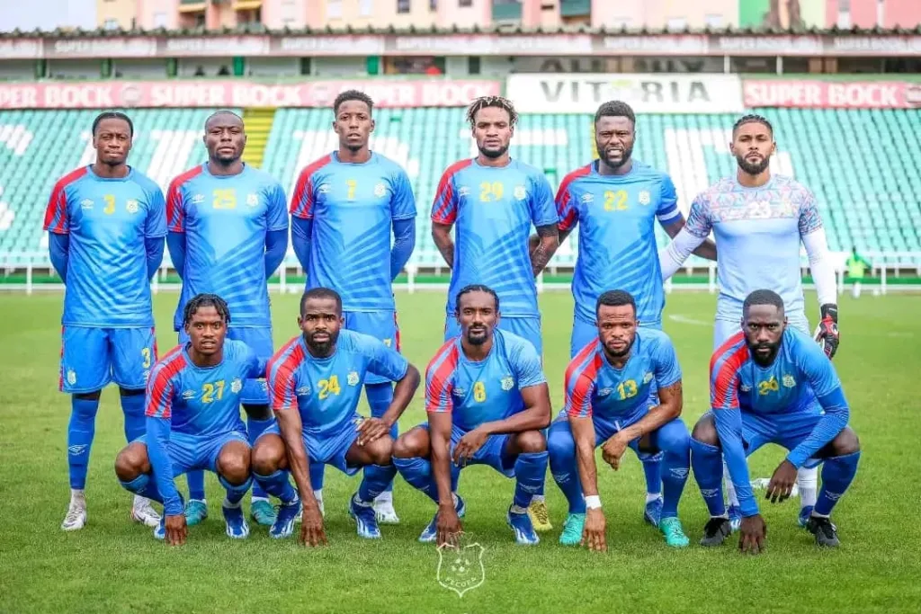 Sur cette photo, l'équipe de football du Léopard RDC est fièrement alignée dans un stade verdoyant. Les joueurs, vêtus de maillots bleus avec des accents rouges, arborent des numéros distincts et des expressions sérieuses, témoignant de leur détermination et de leur esprit d'équipe. Au centre, le gardien de but se démarque avec son maillot coloré, illustrant son rôle unique au sein de l'équipe. Les gradins vides en arrière-plan suggèrent que la photo a été prise avant le début d'un match ou pendant une séance d'entraînement, capturant un moment calme avant la tempête d'activité sur le terrain.